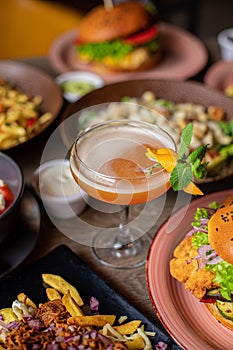 Glass of alcoholic cocktail with mint and orange decor on table full of delicious food. Salad, fries, burgers and drinks