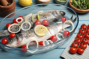 Glass baking tray with sea bass fish and ingredients on light blue wooden table, closeup