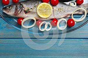 Glass baking tray with sea bass fish and ingredients on light blue wooden table, top view. Space for text