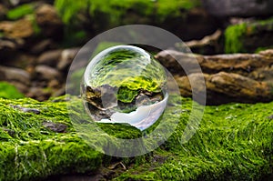 glass ball lens lies on stones covered with green mud and reflects the landscape of the world upside down