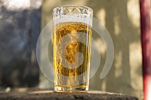 A glass of beer with alcohol. Golden, intoxicating drink in a transparent mug, stands on a wooden stand in the rays of daylight.