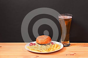 Glass of beer, burger and fried potatoes on wooden table on dark background