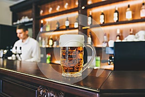 Glass of beer with foam is standing on the wooden bar counter. Old pub with bartender cleaning the dishes on background. Leisure,