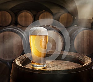 Glass of beer on top of wooden barrel and wood barrels in cellar at the background. Sun ray falling down to it surface.