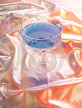 A glass with a blue cocktail drink on a silver background mixed with several different vibrant colors. Futuristic party event
