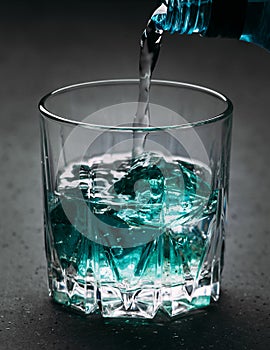 Glass with blue ice cubes. Pouring gin into a glass on a wooden table. Gin in a glass on a black background