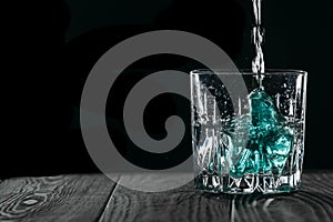 Glass with blue ice cubes. Pouring gin into a glass on a wooden table. Gin in a glass on a black background