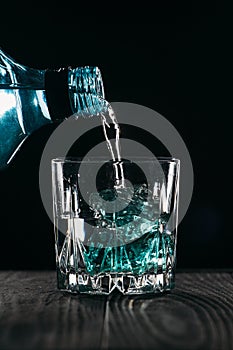 Glass with blue ice cubes. Pouring gin into a glass on a wooden table. Gin in a glass on a black background