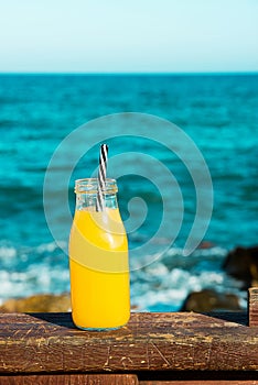 Glass bottle with citrus fruit juice orange with straw on wooden rail. Turquoise sea and blue sky in the background. Tropical