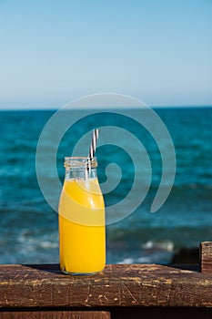 Glass bottle with citrus fruit juice orange tangerine with straw on wooden rail, turquoise sea and blue sky in the background