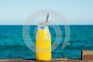 Glass bottle with citrus fruit juice orange tangerine with straw on wooden rail, turquoise sea and blue sky in the background