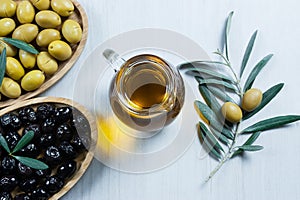 Glass bottle of homemade olive oil and olive tree branch, raw turkish green and black olive seeds and leaves on white table