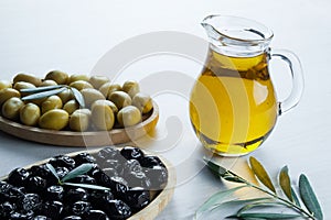 Glass bottle of homemade olive oil and olive tree branch, raw turkish green and black olive seeds and leaves on white table