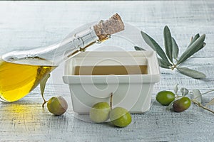 Glass bottle of olive oil over white breakfast square bowl with raw turkish olive seeds and leaves on white rustic vintage table.