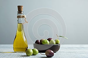 Glass bottle of olive oil and wooden breakfast bowl with raw turkish olive seeds and leaf on white rustic vintage table. Olives b