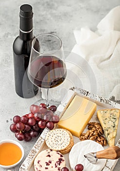 Glass and bottle of red wine with selection of various cheese in wooden box and grapes on light table background. Blue Stilton,