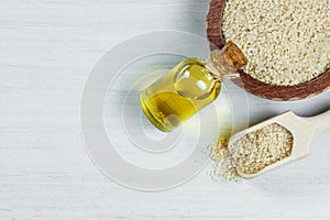 Glass bottle of sesame oil and raw sesame seeds in wooden shovel and bowl on white table