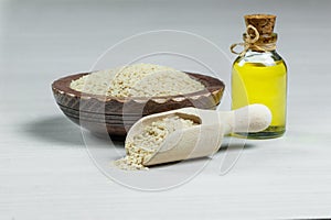 Glass bottle of sesame oil and raw sesame seeds in wooden shovel and bowl on white table