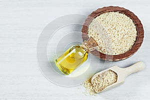 Glass bottle of sesame oil and raw sesame seeds in wooden shovel and bowl on white table