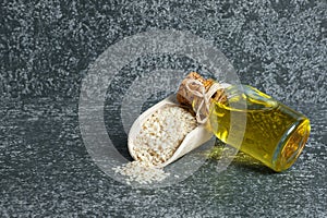 Glass bottle of sesame oil and raw sesame seeds in wooden shovel on grey rustic table