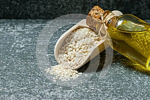 Glass bottle of sesame oil and raw sesame seeds in wooden shovel on grey rustic table