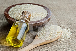 Glass bottle of sesame oil and raw sesame seeds in wooden shovel or spoon and in bowl on burlap sack