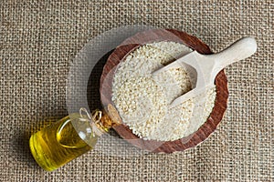 Glass bottle of sesame oil and raw sesame seeds in wooden shovel or spoon and in bowl on burlap sack