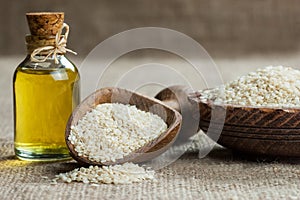 Glass bottle of sesame oil and raw sesame seeds in wooden shovel or spoon and in bowl on burlap sack