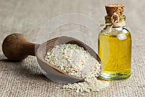 Glass bottle of sesame oil and raw sesame seeds in wooden shovel or spoon on burlap sack