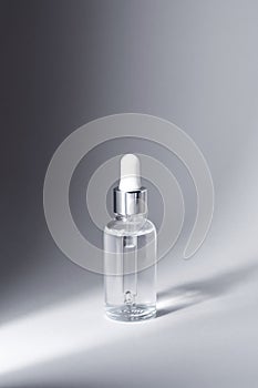 Glass bottle with silver and white pipette in ray of light on grey background mockup. Facial serum in transparent