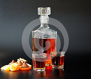 A glass bottle and two glasses of cognac stand on a black background next to sweets, slices and tangerine zest