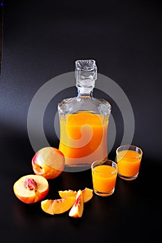 A glass bottle and two glasses of peach liqueur next to ripe fruits on a black background
