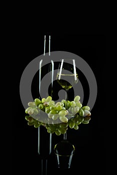 A glass and bottle of white wine with fresh green grapes with reflection on dark