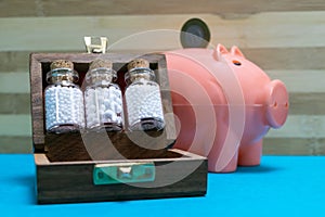 Glass Bottles of homeopathic globules in old wooden box with a blurred coin at pink piggy bank on blue surface and wood background