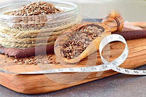 A glass bowl of flax seeds with olive scoop and measuring tape on a olive wood cutting board on grey abstract background