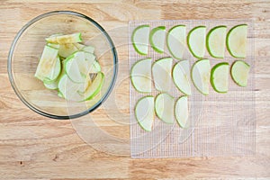 Glass bowl full of granny smith apple slices on a butcher block table, and mesh tray ready for dehydrating apples