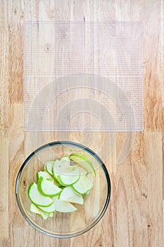 Glass bowl full of granny smith apple slices on a butcher block table, and mesh tray ready for dehydrating apples