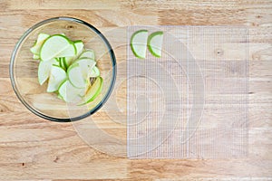 Glass bowl full of granny smith apple slices on a butcher block table, and mesh tray ready for dehydrating apples