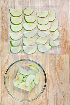 Glass bowl full of granny smith apple slices on a butcher block table, and slices laid out on a mesh tray ready for dehydrating ap