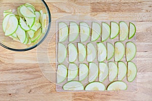 Glass bowl full of granny smith apple slices on a butcher block table, and slices laid out on a mesh tray ready for dehydrating ap