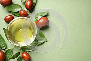 Glass bowl with jojoba oil and seeds on green background, flat lay. Space for text