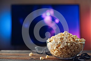 A glass bowl of popcorn, 3d glasses and remote control in the background the TV works. Evening cozy watching a movie or TV series