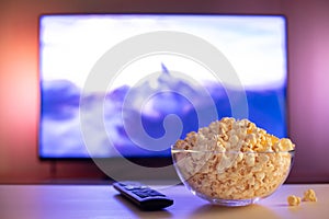 A glass bowl of popcorn and remote control in the background the TV works. Evening cozy watching a movie or TV series at home