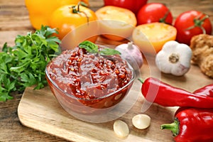 Glass bowl of tasty adjika and ingredients on wooden table