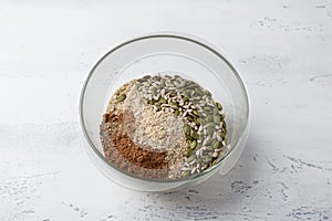 Glass bowl with whole sunflower and pumpkin seeds, ground seeds, cocoa and spices on a light gray table, top view. Stage