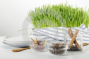 Glass bowls full of spices, plates, wooden spoons, fresh green plants on the white kithcen table against bright wall