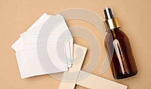Glass brown bottle with cosmetic spray and paper business cards on brown background, top view