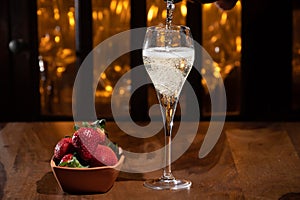 Glass of brut champagne bubbles wine in tulip glass and bowl with fresh strawberries on evening bar lights background