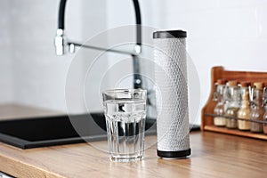A glass of clean fresh water and carbon filter cartridge on wooden table in a kitchen interior. Installation of reverse