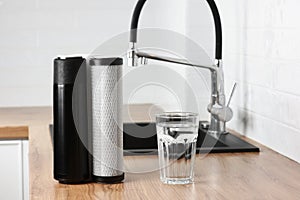 A glass of clean fresh water and set of filter cartridges on wooden table in a kitchen interior. Installation of reverse